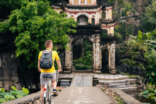 Tourist riding bicycle on stone bridge towards historic entrance gate of Bich Dong Pagoda in Ninh Binh Vietnam. Cultural travel experience, Asian heritage and active holiday concept.