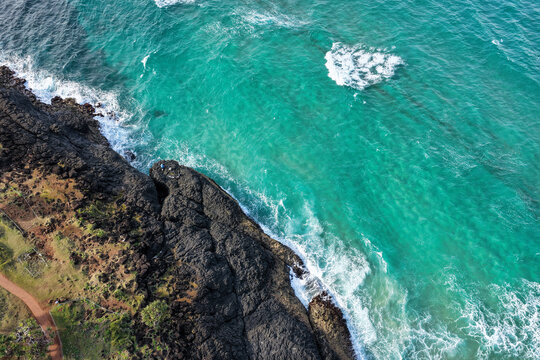 Aerial view of Fingal Head featuring dark volcanic basalt rock formations and turquoise ocean waves crashing against the coastline in Fingal Head, New South Wales, Australia.