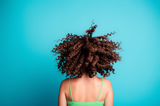 Young woman with curly hair flipping her head in a vibrant turquoise studio background, back view showing motion, texture and playful summer style