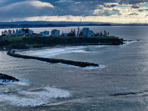 Aerial view of Duranbah Beach with its rock breakwater extending into the ocean, surrounded by coastal buildings and lush headlands under a dramatic sky in Duranbah, New South Wales, Australia.
