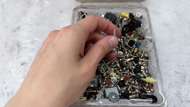 A hand picks a small screw from a clear plastic box full of screws, springs and clips. Device repair, spare parts, metal hardware, loose fasteners
