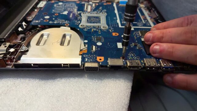 A male hand uses a screwdriver on a blue laptop motherboard with ports on white foam. Computer repair, device service, spare parts, PC hardware