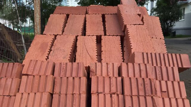 Rows of red ceramic bricks are stacked in a pile outdoors near a building. Construction materials, building supplies, masonry work, home renovation