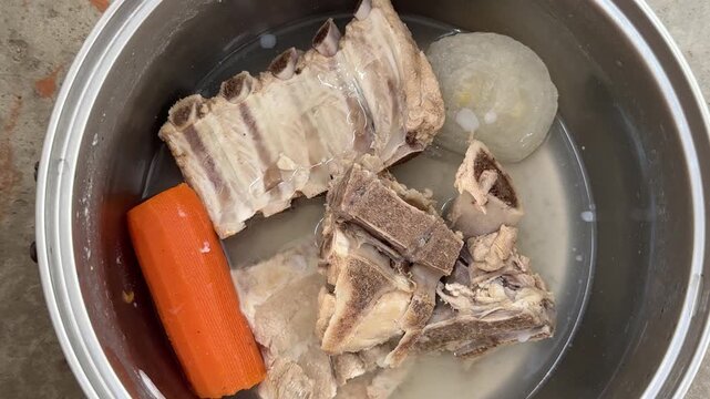 Boiled pork bones with a carrot and onion sit in water inside a steel pot. Bone broth, soup preparation, home cooking, meat stock