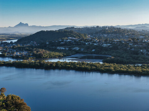 Aerial view of the Tweed River winding through Chinderah with lush mangroves, residential areas, and distant mountains under a clear morning sky in Chinderah, New South Wales, Australia.
