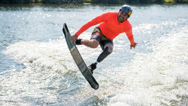 Joyful adaptive man with leg prosthetic wakeboarding over lake wake doing grab trick