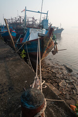 Weathered blue wooden fishing boats tied to a rusted mooring bollard with thick ropes at a calm harbor during a hazy morning sunrise © Zaenal