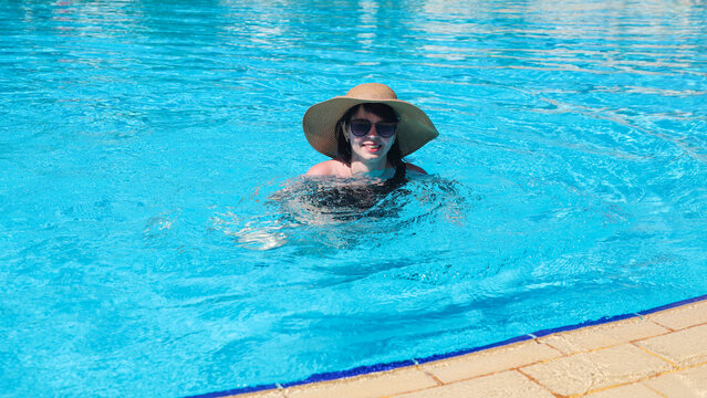 Woman in sunglasses and wide brimmed hat swimming leisurely sunny pool.