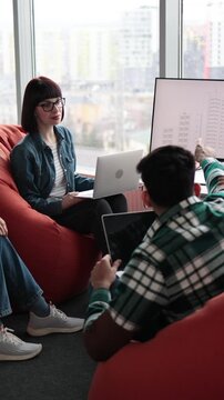 A group of four diverse colleagues are engaged in a discussion while working on laptops and a monitor in a relaxed, modern office setting with large windows overlooking a city
