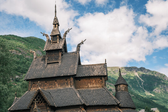 View of Borgund Stave Church with its intricate wooden architecture, shingled roof, and dragon carvings set against green mountains and a blue sky in Borgund, Vestland, Norway.