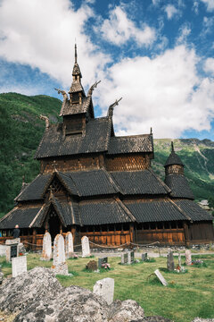 View of the historic Borgund Stave Church featuring intricate wooden carvings and dragon-head roof decorations, surrounded by a cemetery against a mountainous landscape in Borgund, Vestland, Norway.