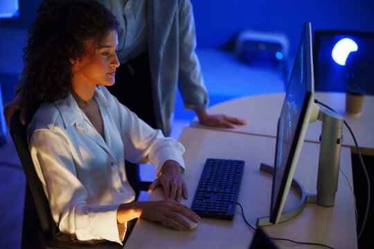 Female cybersecurity analyst monitoring computer systems with a colleague in a blue-lit operations center, representing network security, threat detection, data analysis and digital protection.