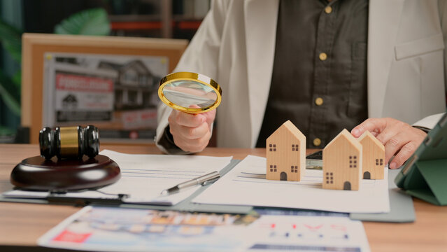 Businessperson holding magnifying glass examining wooden house model paperwork legal document gavel desk mortgage loan property purchase investment planning strategy asset risk assessment.