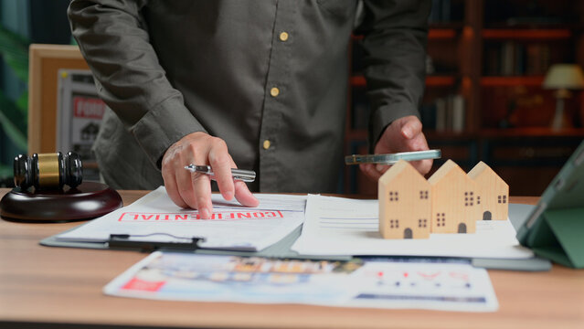 Lawyer reviewing confidential property contract documents holding pen smartphone wooden house model desk gavel arbitration court judge real estate transaction closing agreement signature plan.