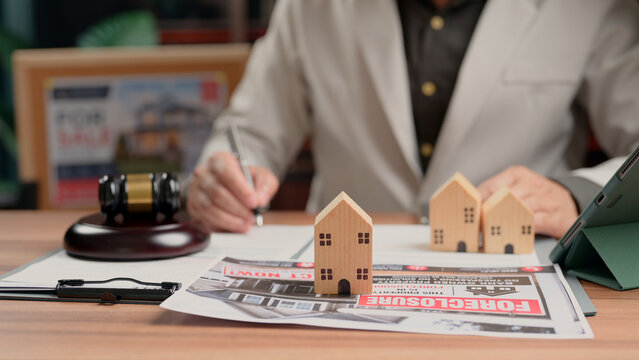 Person wearing jacket signing foreclosure document contract desk miniature wooden house model legal gavel tablet computer corporate office workspace business property management.