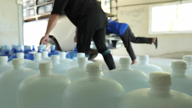 Rows of drinking water gallon bottles at a water bottling factory, with a worker in the background loading gallons onto a pickup truck for delivery and distribution.
