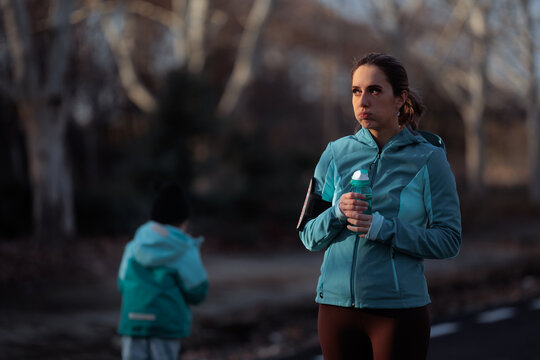 Tired Mother Holding Water Bottle Taking Break From Outdoor Workout. Active mom resting during fitness training while child plays 
