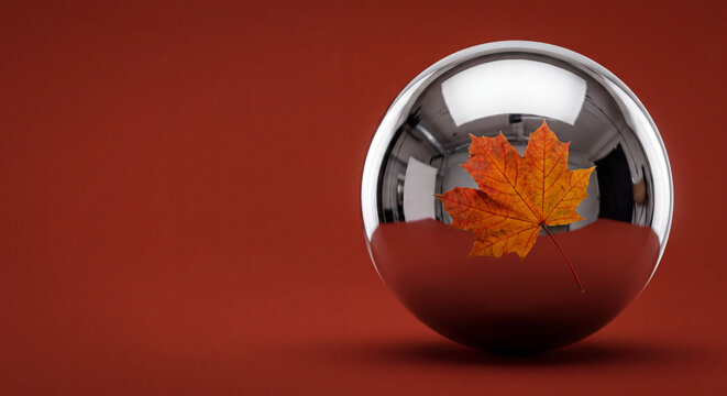 Chrome sphere reflecting an orange maple leaf on a red background, autumn concept