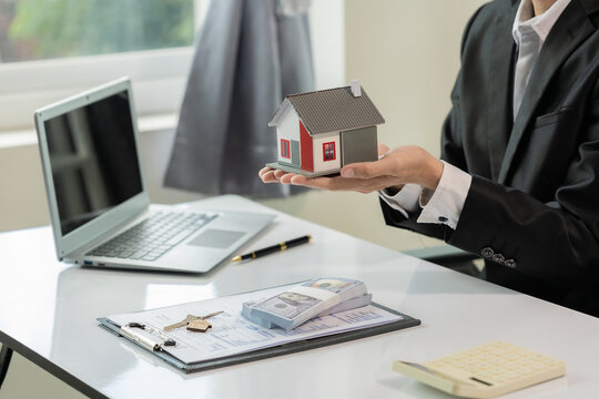 Real Estate Agent Holding House Model with Cash Stack and House Keys on Desk