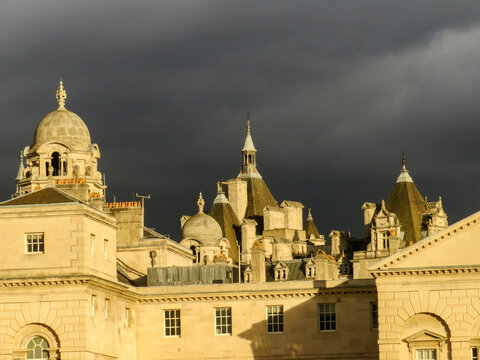 View of the historic Horse Guards building with its distinctive domes and towers illuminated by warm sunlight against a dramatic dark stormy sky in London, England, United Kingdom.
