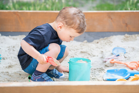 Cute toddler digging in the sand with a toy scoop in a backyard sandbox.