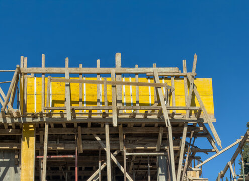 Weathered timber formwork frame an unfinished concrete building beneath a vivid blue sky