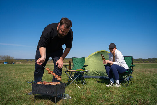 Couple enjoying outdoor camping, with a man grilling shish kebab over a portable barbecue and a woman relaxing in a chair, drinking a bottle of beer on a sunny day in a green field
