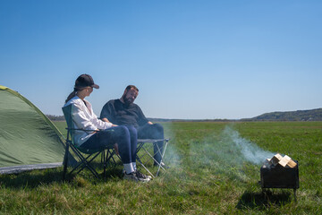 Fototapeta premium Couple relaxing in camping chairs by a green tent, enjoying a small wood-burning campfire under a clear blue sky in an open grassy field, peaceful summertime escape