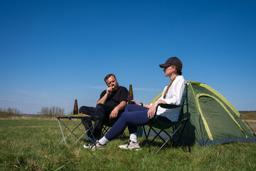 Fototapeta premium Two people are relaxing in camping chairs next to a tent in a green field under a clear blue sky, enjoying a snack and drinks during their outdoor leisure time