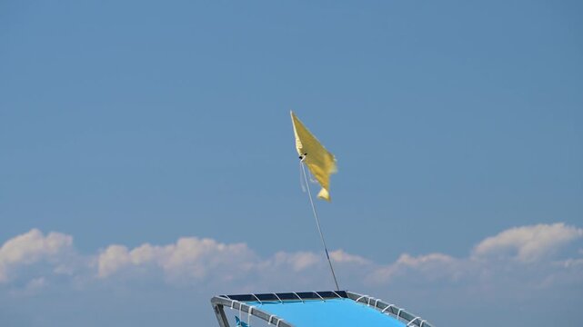 A small yellow warning flag flutters in the wind against a bright blue sky with white clouds above a blue boat canopy.