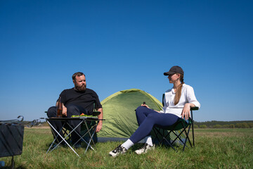 Fototapeta premium Couple resting and enjoying drinks in camping chairs next to their tent in a sunny green field, symbolizing leisure, outdoor activity, and a shared travel experience
