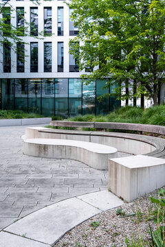 Curved concrete benches trace a courtyard where landscaping and trees soften modern glass architecture, as an urban walkway bends around the planted edge.