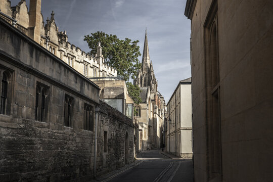 View of a narrow historic street featuring ancient stone architecture, a prominent church spire, and weathered masonry walls under a soft sky in Oxford, England, United Kingdom.