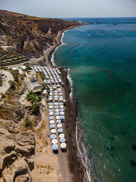 Aerial view of the volcanic black sand beach with rows of white umbrellas, turquoise sea, and rugged cliffs in Fira, Greece.