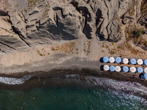Aerial view of a volcanic black sand beach with rows of umbrellas and turquoise water at the base of steep cliffs in Fira, Greece.