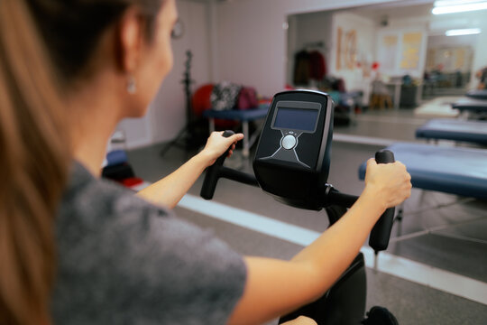 Female Patient Training On Recumbent Bike At Rehabilitation Center. Over the shoulder view of young woman exercising on stationary bicycle in clinic
