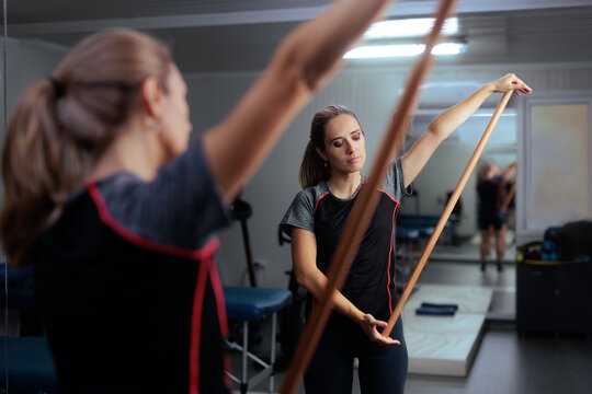 Focused Patient Exercising With Wooden Stick At Rehabilitation Center. Young woman using gymnastic wooden pole for mobility training in clinic mirror
