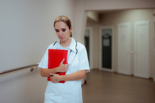 Sad Female Doctor Holding Red Clipboard In Hospital Corridor. Disappointed young nurse with stethoscope clutching medical folder in clinic hallway