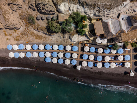 Aerial view of Fira beach with rows of blue and white umbrellas on dark volcanic sand, turquoise sea, and rocky cliffs in Fira, Greece.