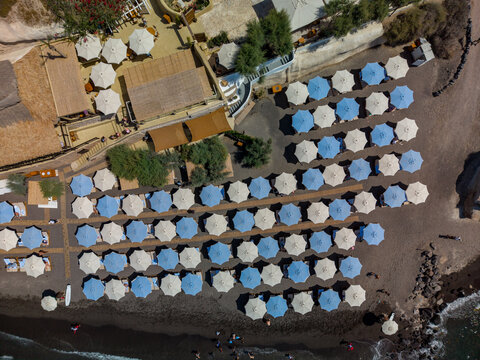 Aerial view of rows of blue and white umbrellas on the dark volcanic sand of the beach with a restaurant terrace in Fira, Greece.