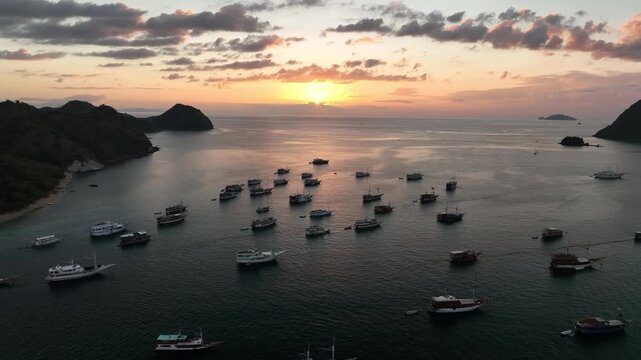 Aerial view of numerous traditional wooden boats anchored in the calm waters of Labuan Bajo bay during a golden sunset in Komodo, East Nusa Tenggara, Indonesia.