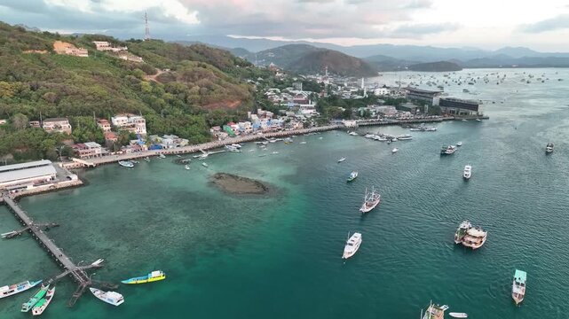 Aerial view of Labuan Bajo harbor with traditional wooden boats anchored in the turquoise bay, surrounded by lush green hills and coastal buildings in Komodo, East Nusa Tenggara, Indonesia.