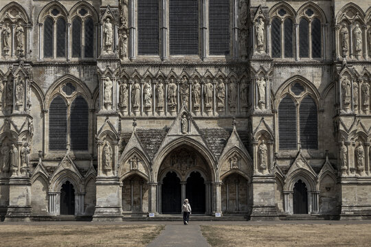View of the intricate Gothic facade and stone sculptures of Salisbury Cathedral with a person walking towards the entrance in Salisbury, England, United Kingdom.
