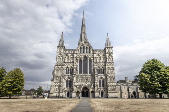 View of Salisbury Cathedral with its iconic spire, intricate Gothic stone facade, and surrounding green lawn under a cloudy sky in Salisbury, England, United Kingdom.
