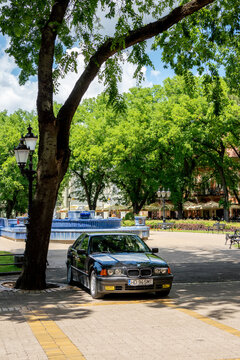 Black BMW 3 Series E36 sedan parked beneath mature trees beside a blue fountain (plava fontana) in center of Subotica, Serbia, June 7, 2026. Vertical image.