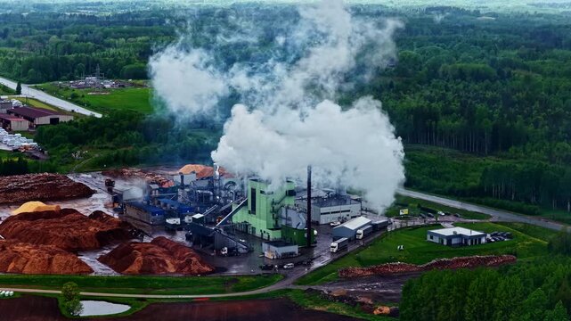 Aerial view of wood processing factory emitting smoke with large piles of wood chips and surrounding forest in Latvia.