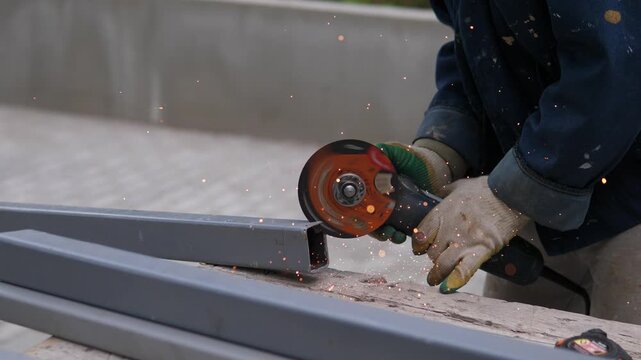 A focused worker wearing gloves and protective clothing is cutting a metal beam with a grinder at a construction site. The industrious individual demonstrates precision and skill in their t.
