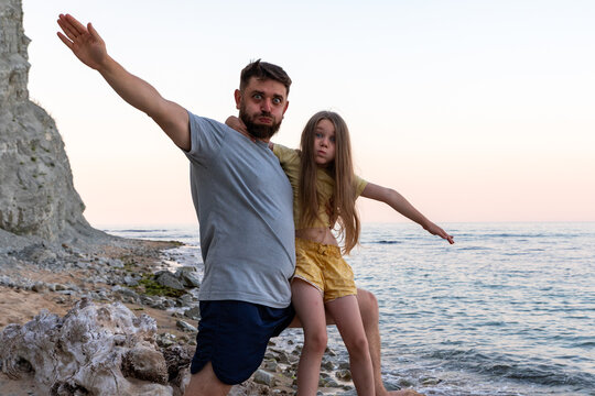 Father and daughter posing together on a rocky beach at sunset, arms outstretched, enjoying a playful and joyful moment by the water.