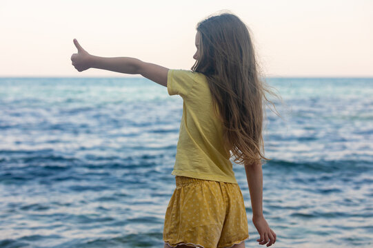 Young girl in yellow standing by the sea, giving a thumbs up gesture, symbolizing positivity and fun summer days.
