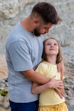Loving father hugging his smiling daughter outside on a rocky beach, showing family connection, warmth, and happiness in a natural setting.